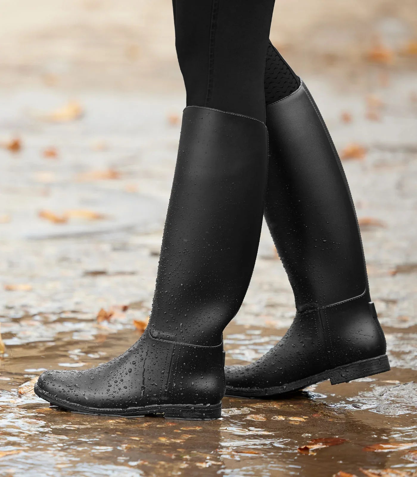 Child wearing long black waterproof riding boots, standing in a shallow puddle with autumn leaves on the ground.